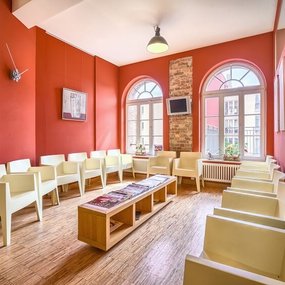 Modernes Wartezimmer mit roten Wänden, weißen Stühlen und großen Fenstern im Tageslicht.