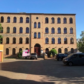 Backstein-Gebäude mit Schriftzug Drahtzaun-Fabrik, mehrere Autos parken davor.